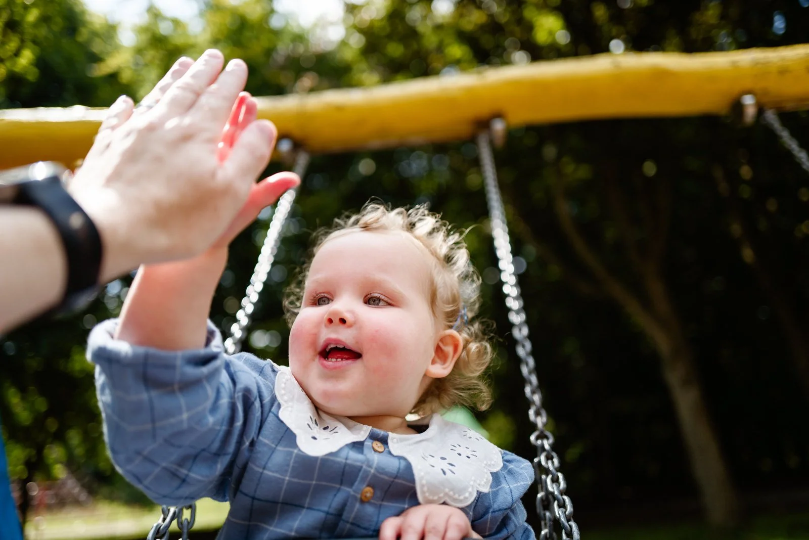 A young girl on a swing reaching out to high-five an adult's hand in a park.
