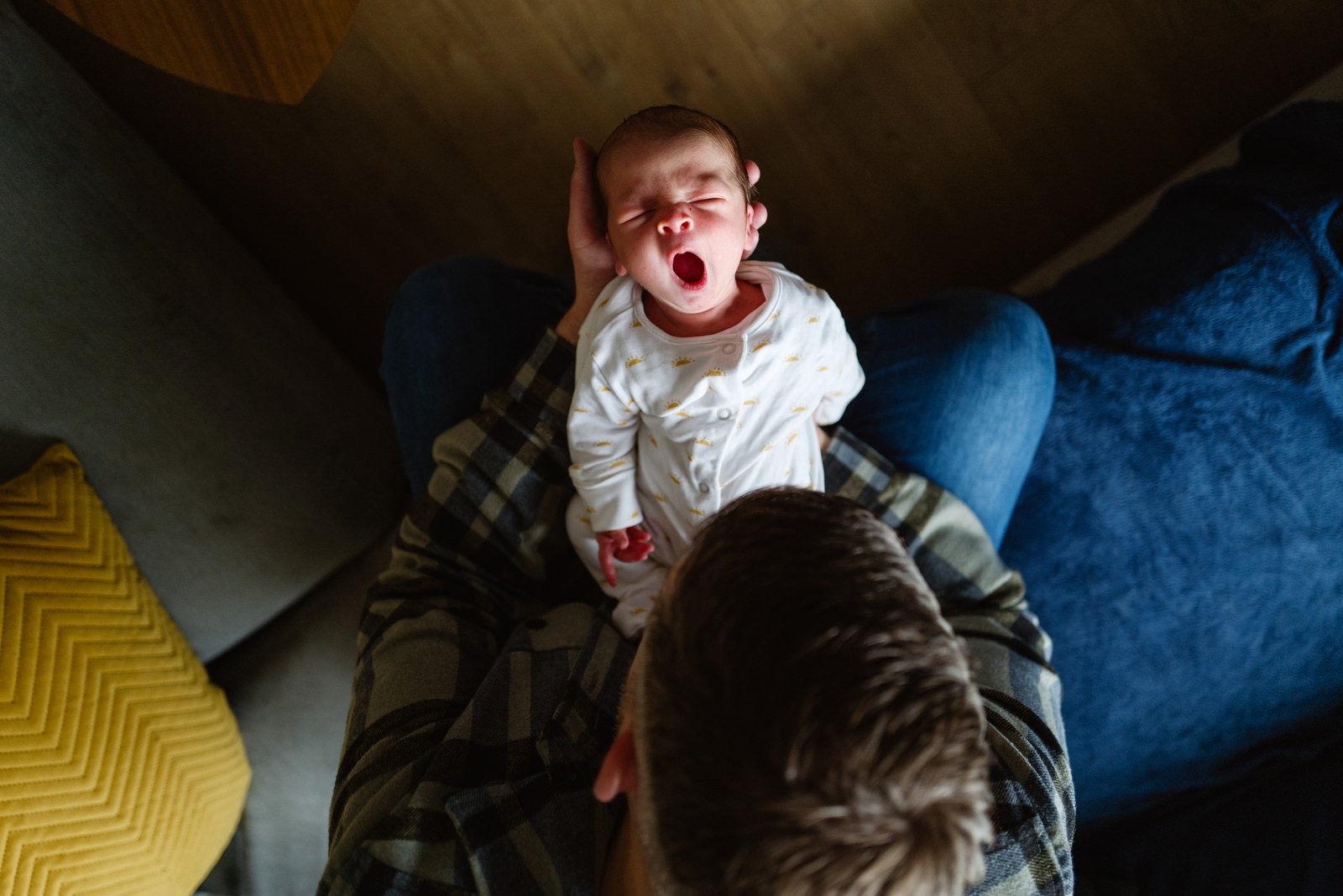 A yawning baby sitting on an adult's lap indoors, shot from above.