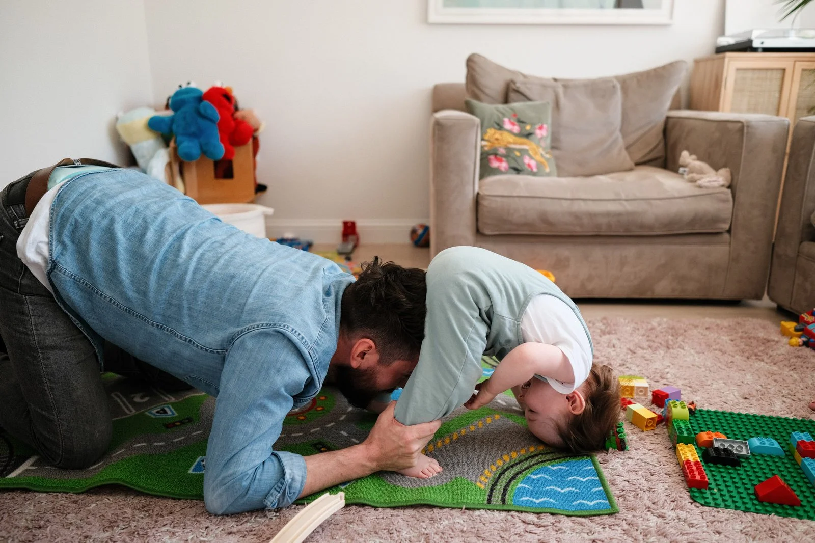 A father and child playfully engaging on a colorful play mat in a living room, with toys scattered around and a beige couch in the background.