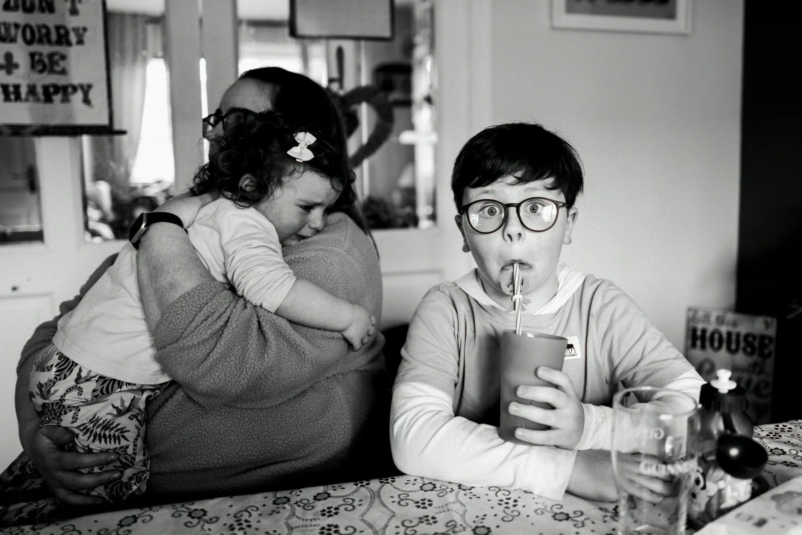 A boy wearing glasses is sitting at a table, sipping from a cup with a straw, while a young girl is hugging an adult woman, who is partially obscured, in the background.