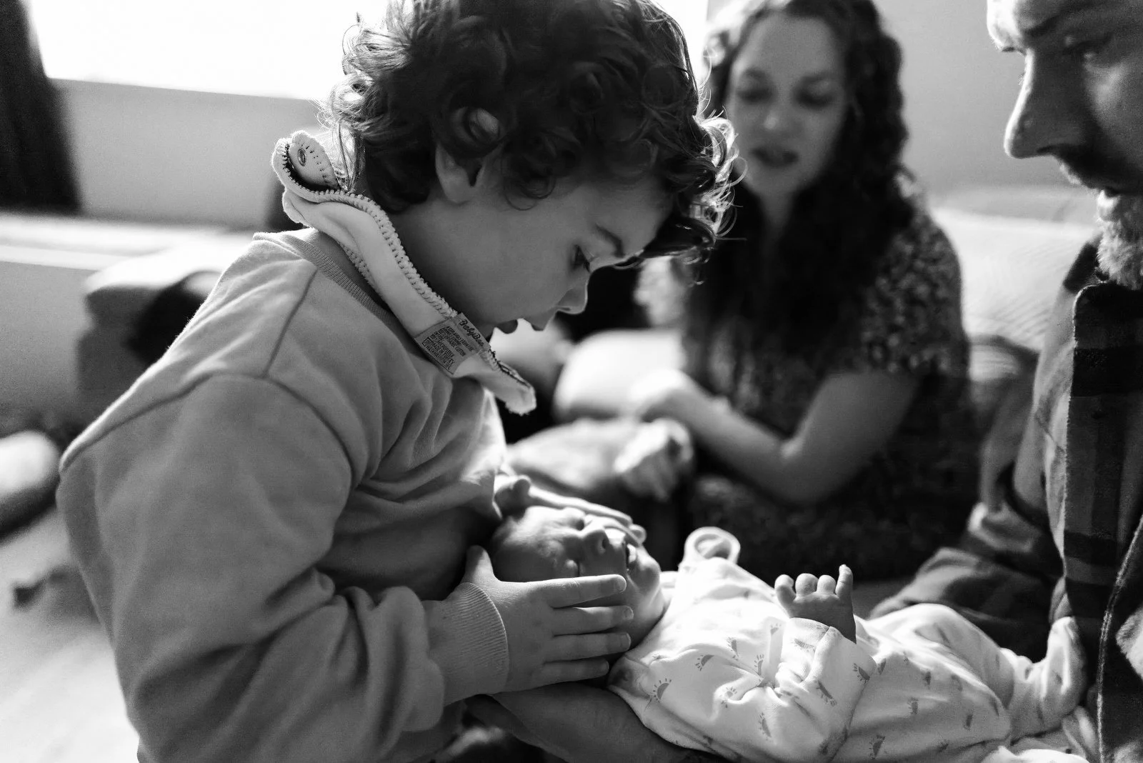 A young child with curly hair gently touches a newborn baby lying on a parent's lap. A woman and a man look on, with the woman speaking, in a cozy indoor setting.