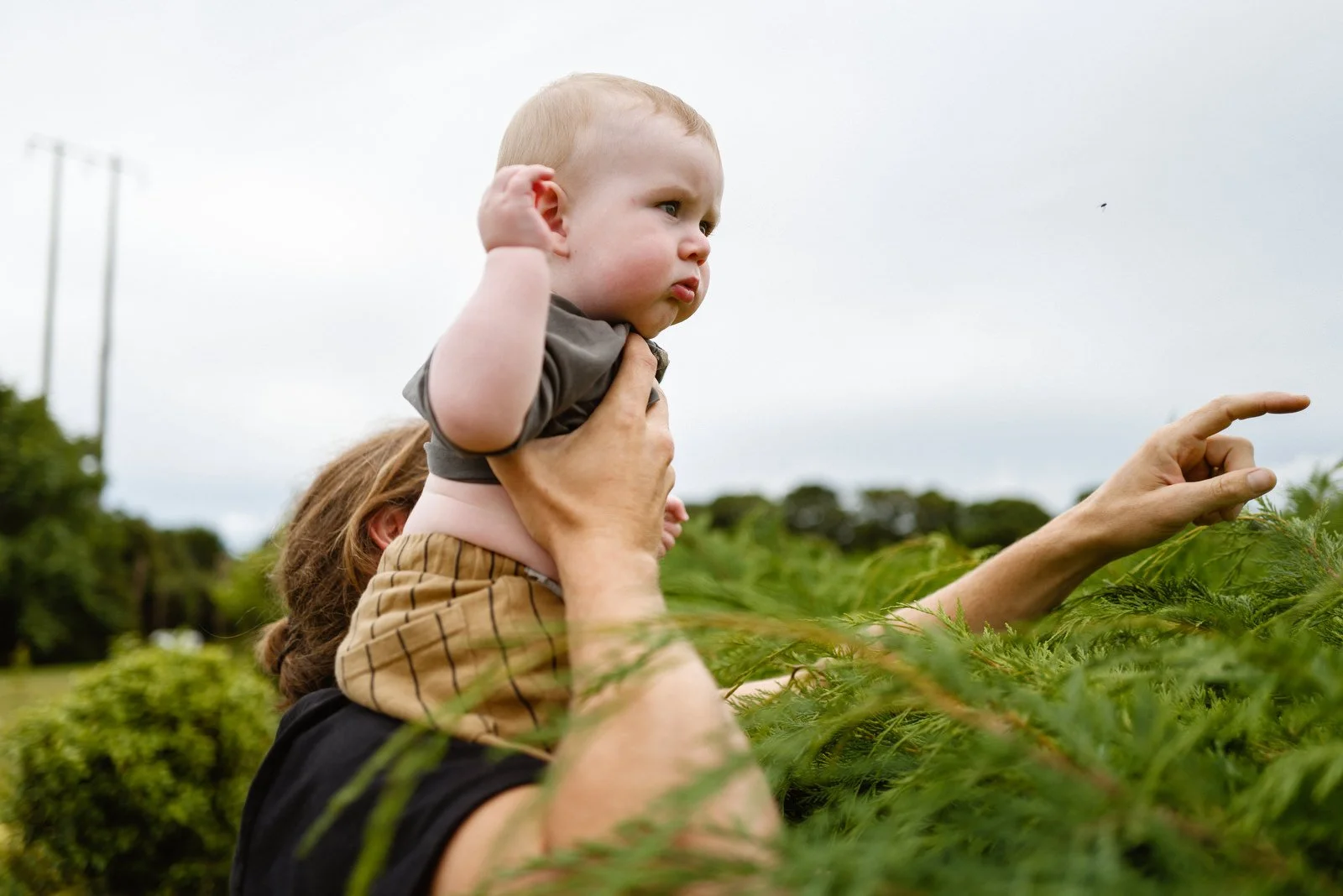 A woman giving a young child a piggyback ride outdoors, with the child pointing at something in the distance. They are surrounded by greenery and cloudy skies.