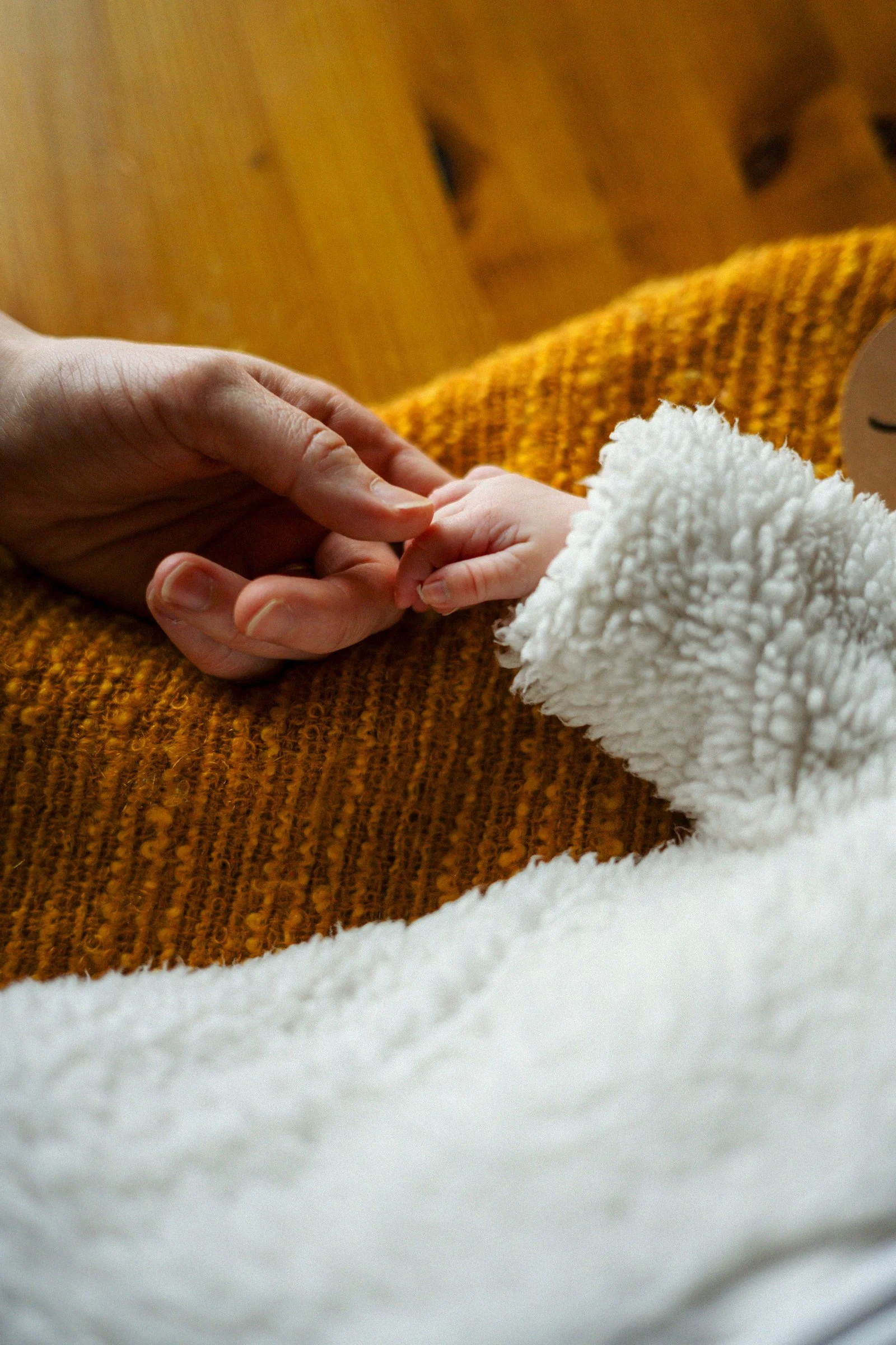A close-up of a person's hand gently holding a baby's hand, with the baby wearing a fluffy white garment. The background includes a wooden surface and a orange-brown blanket.