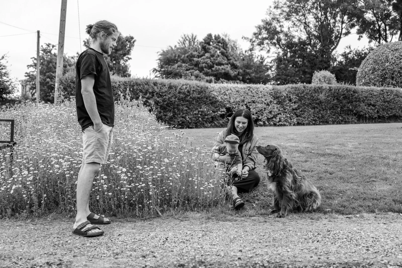 A woman kneeling on the grass holding a baby and talking to a dog while a man stands nearby looking on in a park with bushes and trees in the background.