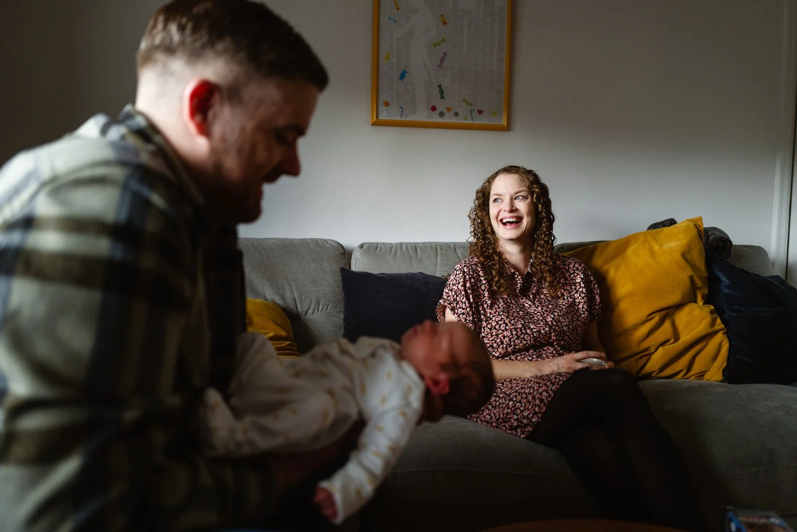 A man holding a newborn baby and hugging a woman sitting on a couch in a living room, all smiling and sharing a joyful moment.