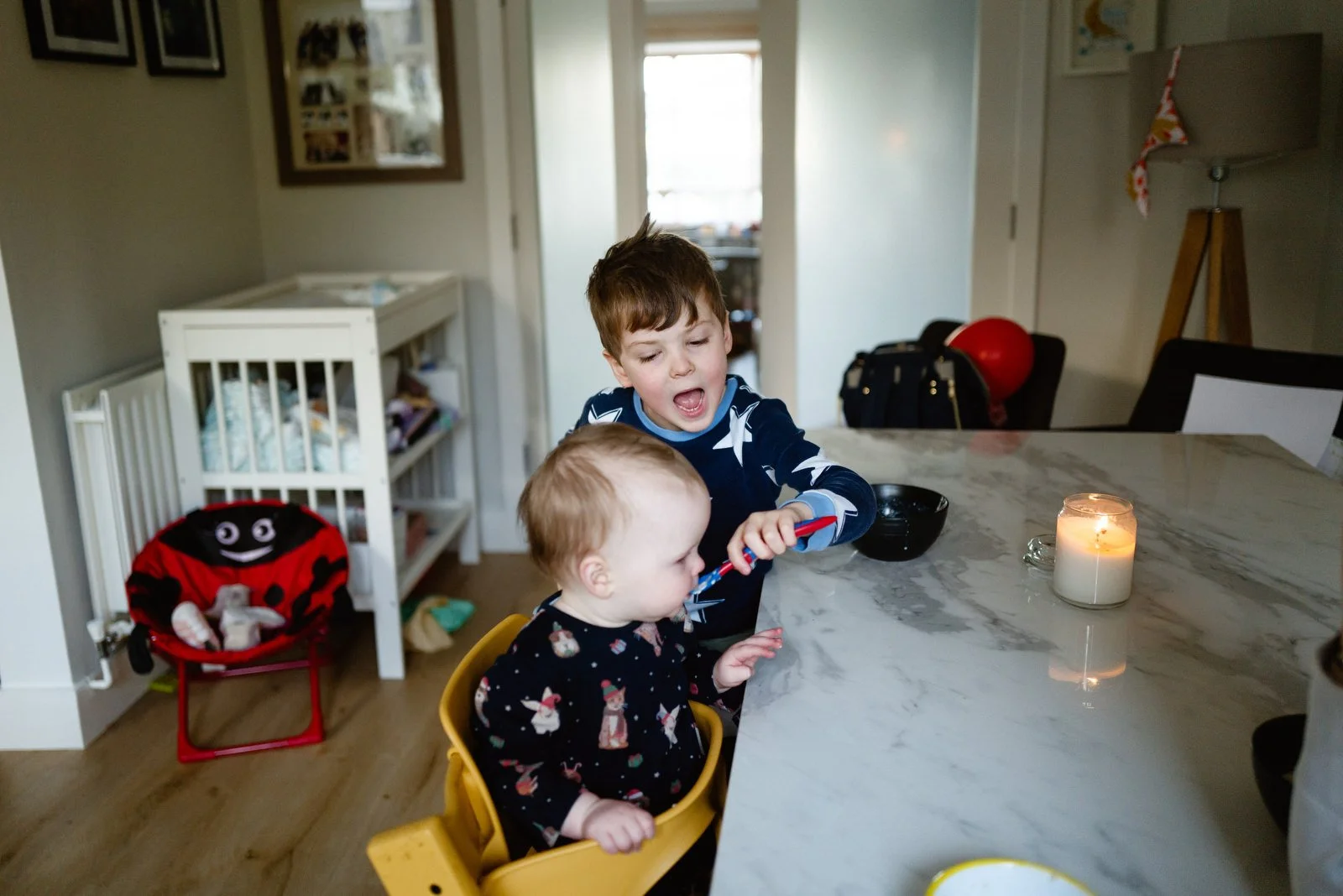 A boy and a toddler sitting at a kitchen island, the boy appears to be brushing the toddler's teeth. There is a candle on the table, and a stuffed ladybug chair in the background.