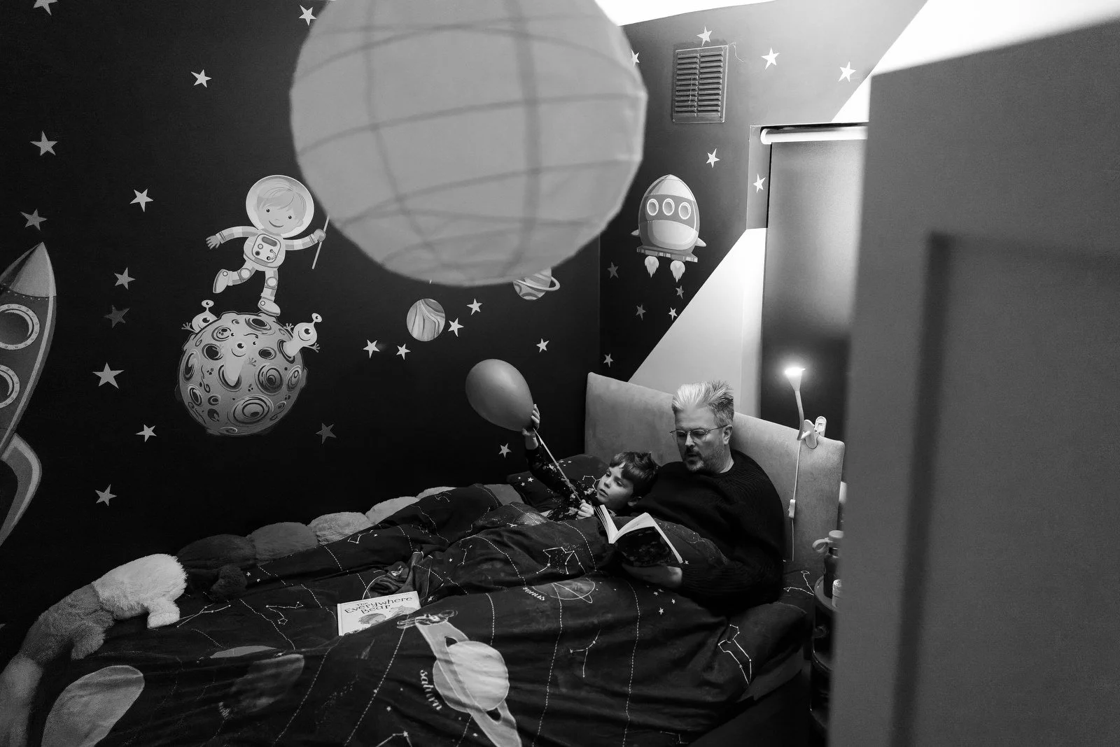 A child and an adult lying in bed with a space-themed mural on the wall behind them, featuring planets, stars, a rocket, a spaceship, and an astronaut.