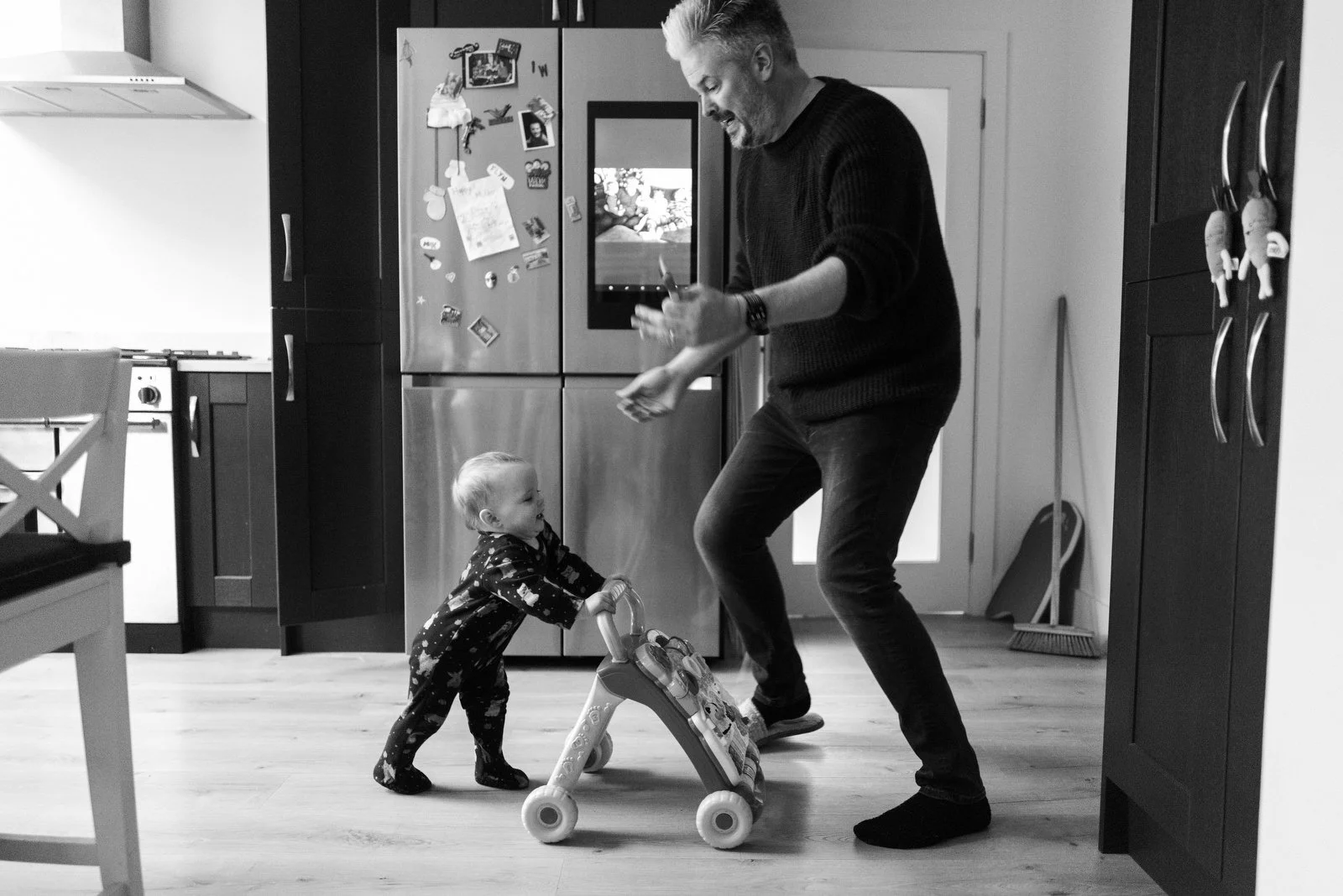 A man and a toddler playing with a toy walker in a kitchen. The man is smiling and holding an object, while the toddler is pushing the toy and looking up at the man.