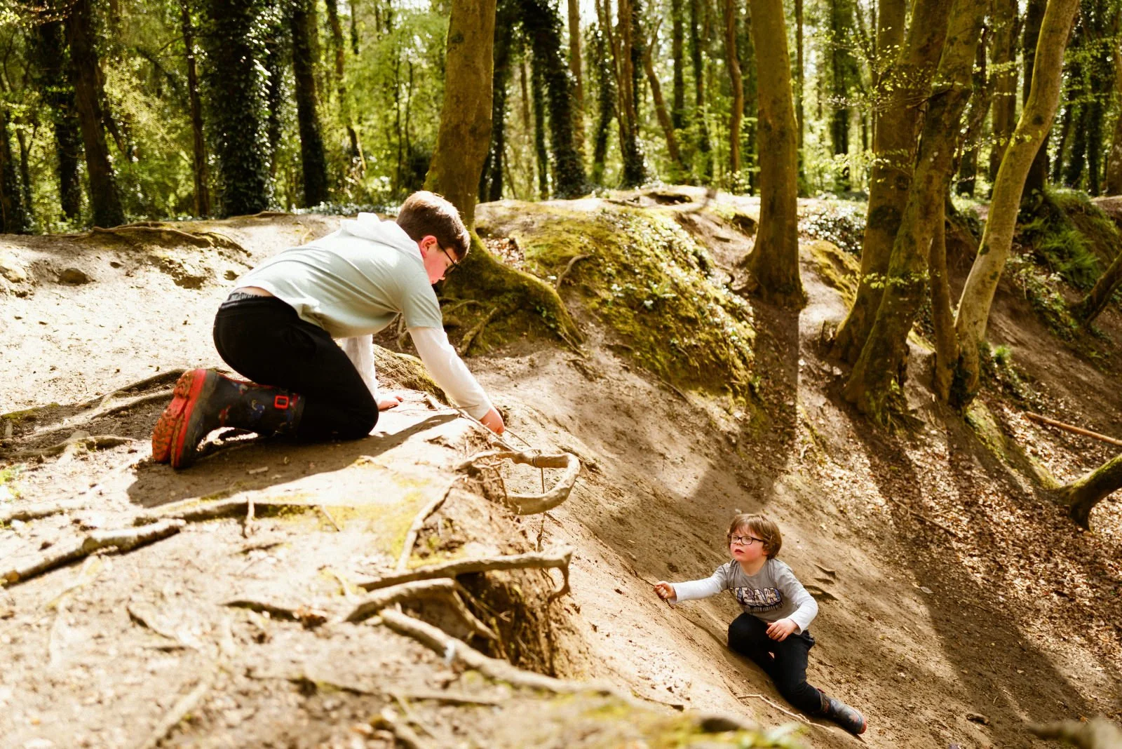 A man helping a young boy climb up a steep, sandy hill in a forest with tall trees and sunlight filtering through the leaves.