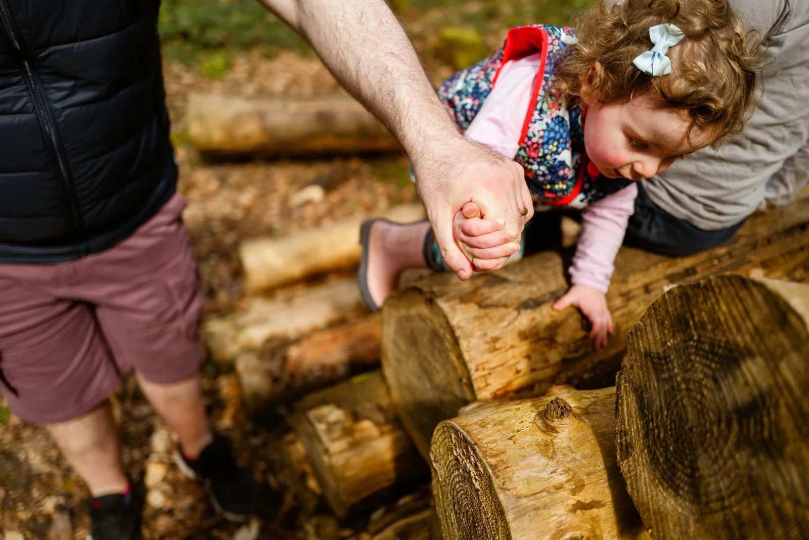 A young girl with curly hair and a bow in her hair leaning over a log, holding onto an adult's hand for support while on a log balance beam outdoors.