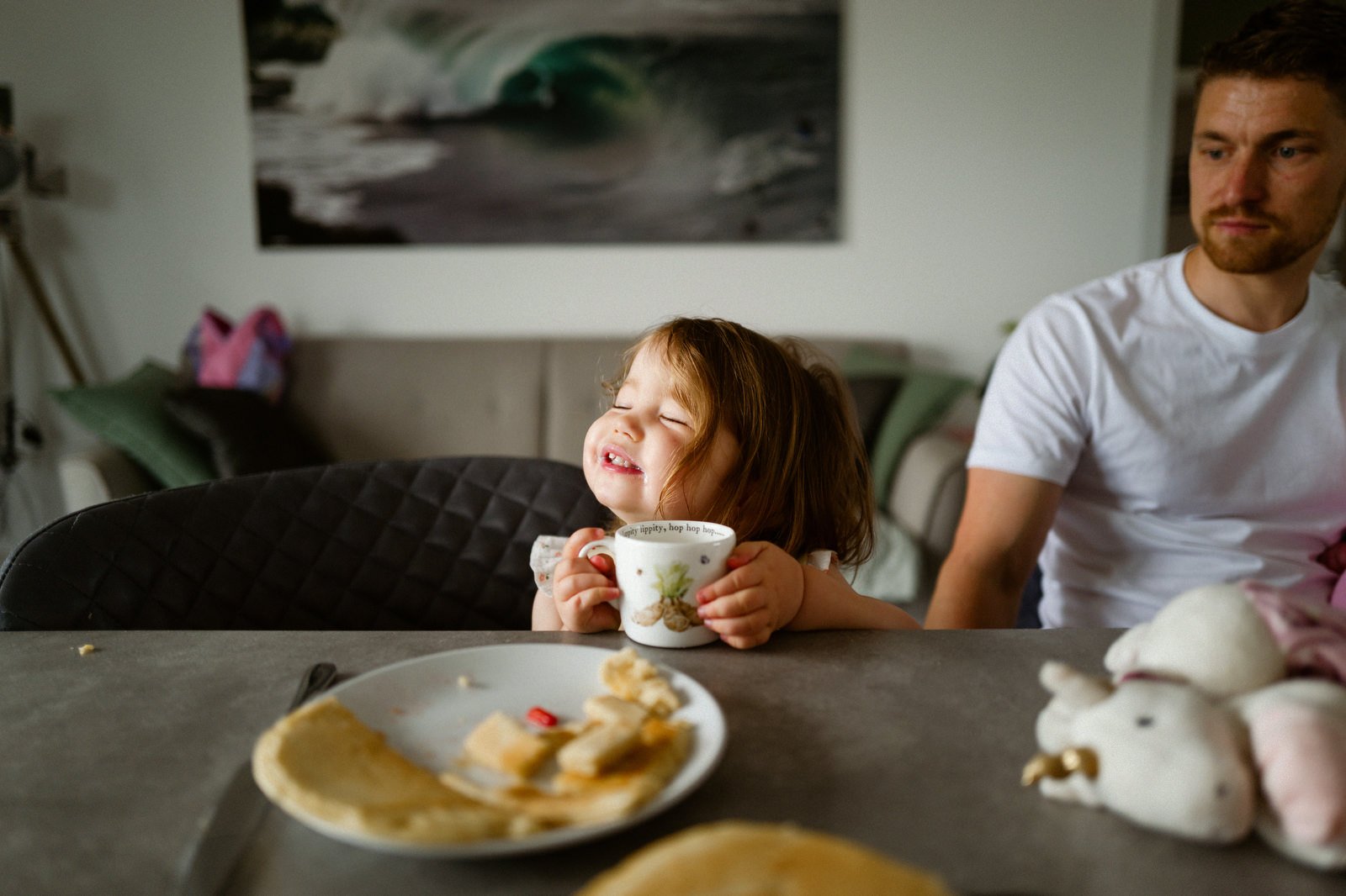 A young girl with curly hair smiling and holding a mug, sitting at a dining table with a plate of bread and a knife, and a man in a white t-shirt sitting nearby in a living room with a painting of a wave on the wall.