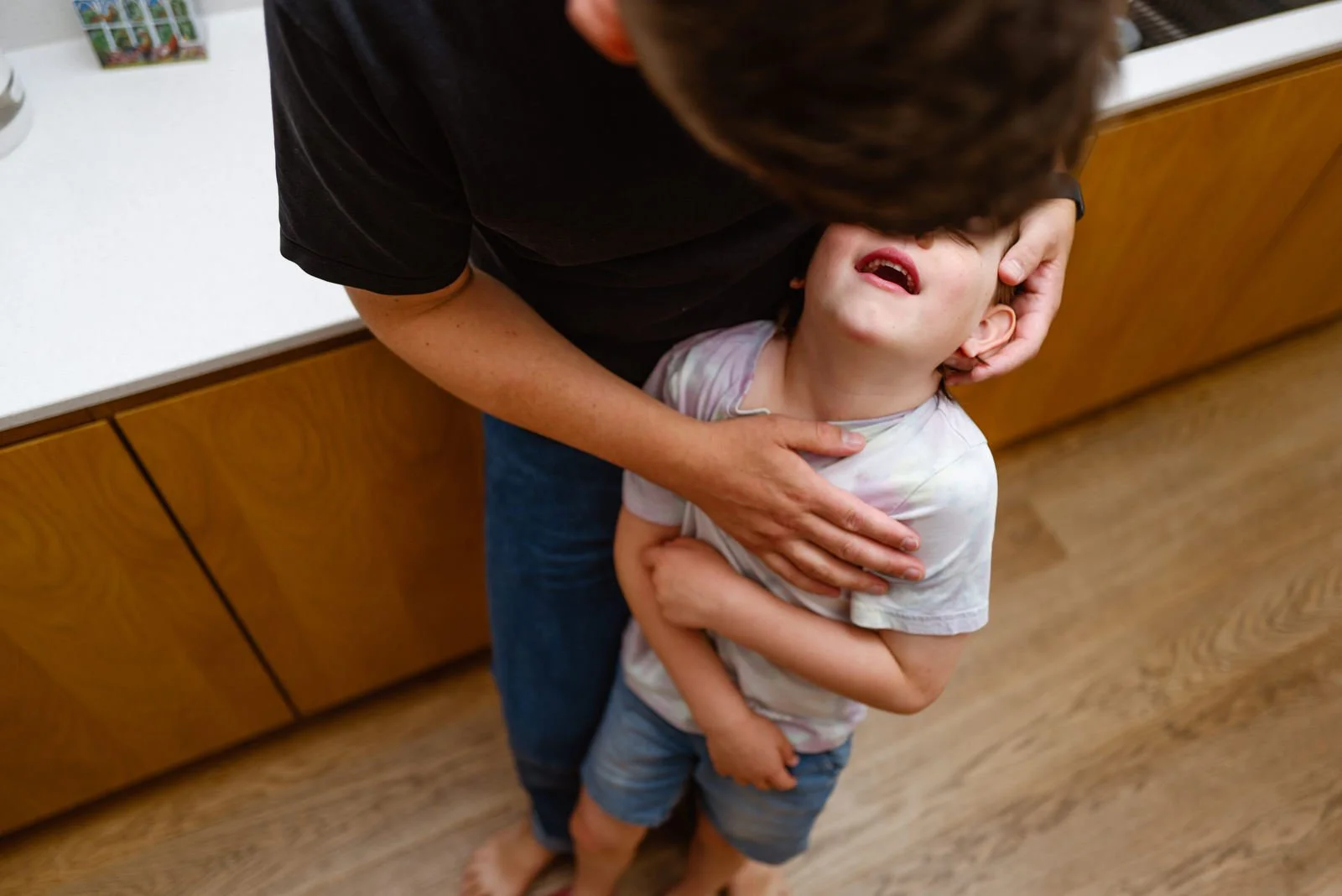 A young child appears to be crying or distressed, being comforted by an adult who is holding the child's head and shoulder. The scene is indoors, with wooden cabinets and a white countertop in the background.