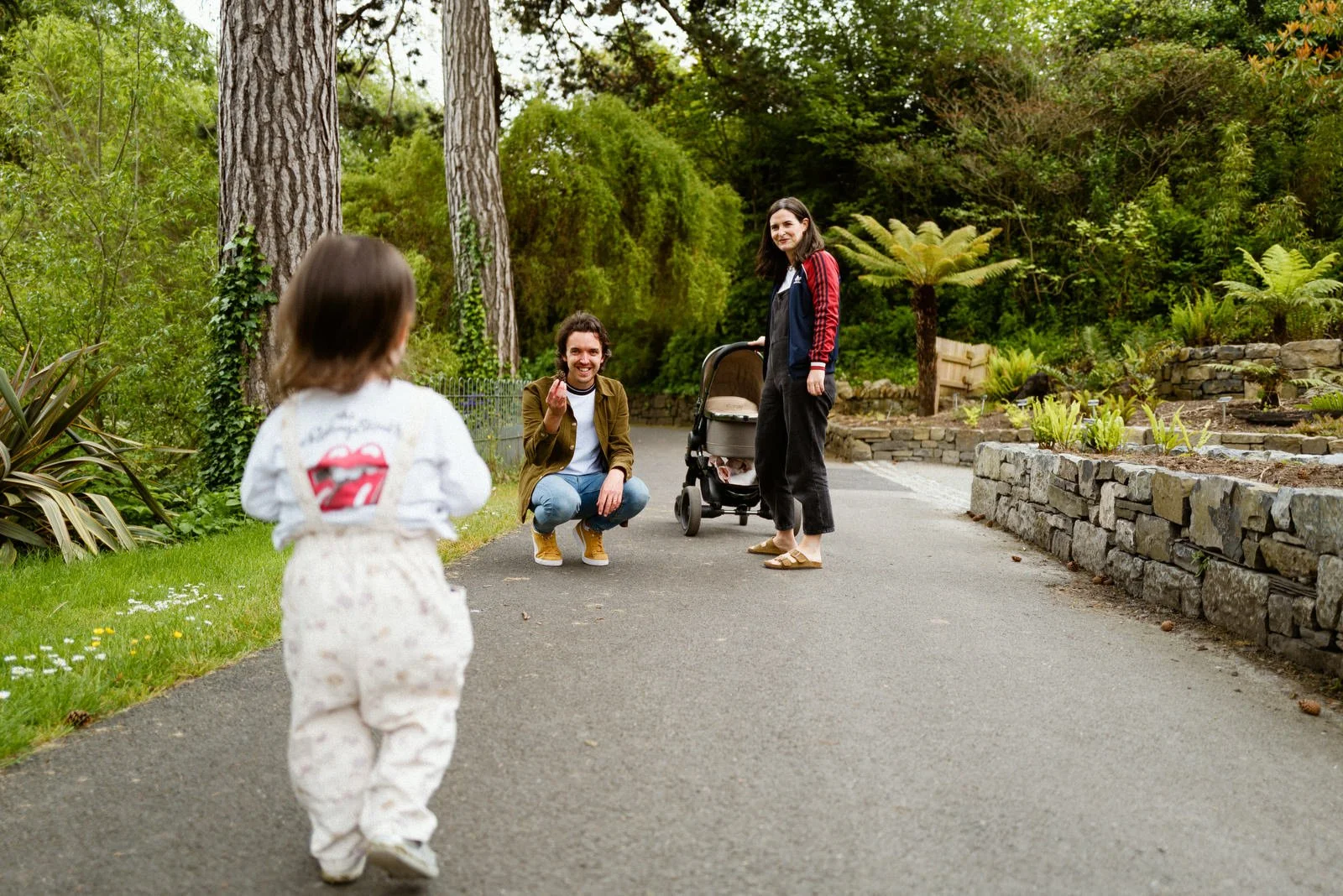 A woman and man interacting with a small girl on a park pathway, with a woman standing near a stroller and trees and plants in the background.