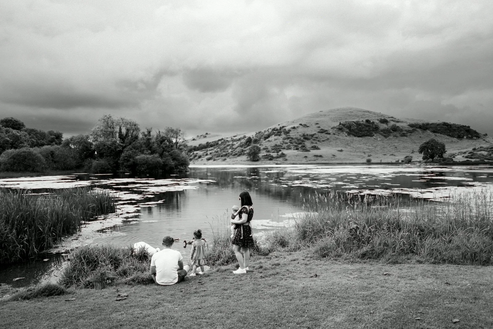 Black and white photo of a family by a lake with a hill in the background. The family includes a man, a woman holding a child, and a young girl, all near the water's edge, with cloudy skies overhead.