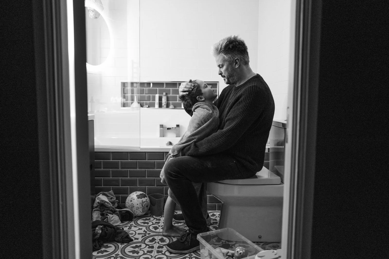 A man and a boy sitting on a toilet in a bathroom, with the man gently holding the boy's head, sharing an intimate moment.