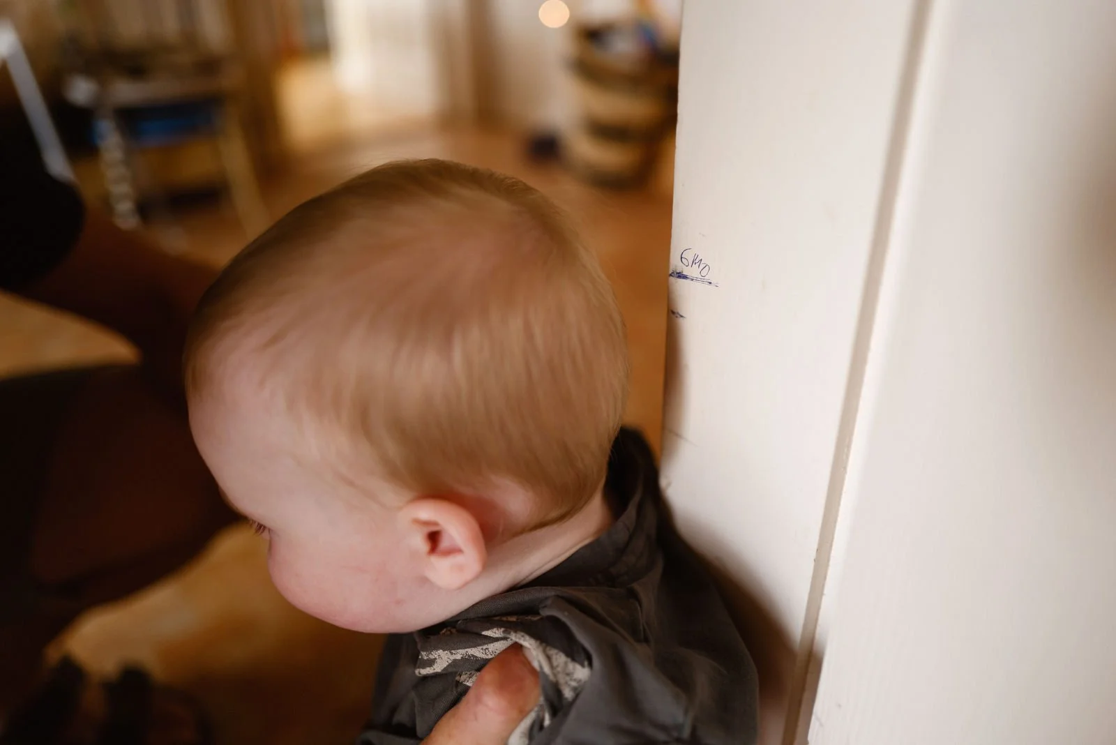 A young child with light brown hair facing a white wall, standing on a wooden floor in a warmly lit room.