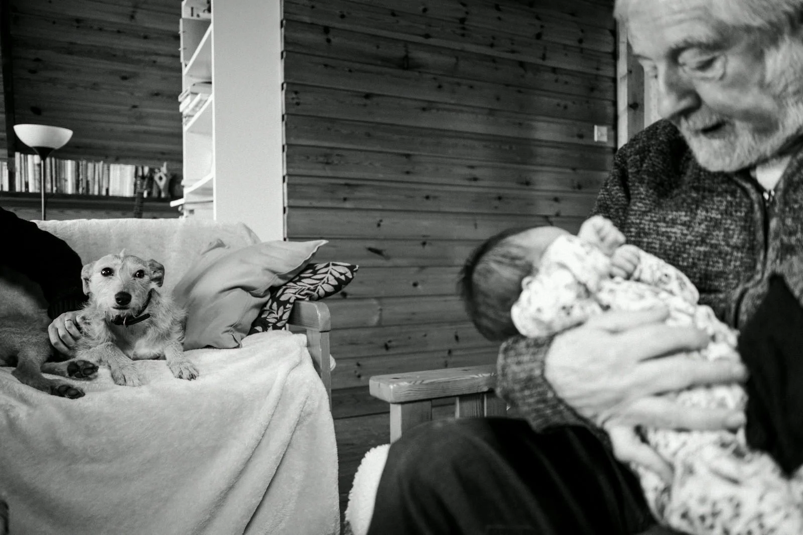 A man smiling and holding a newborn baby wrapped in a blanket, while a dog lies on the sofa looking at the camera. The setting appears to be a cozy room with wooden walls.