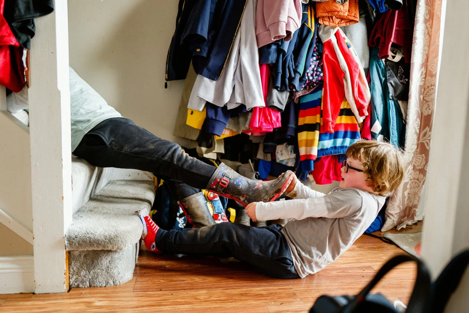 A young boy with glasses and curly hair lies on the floor, holding his feet up with his legs on a nearby sofa. An adult's legs are visible on the sofa. The background shows a closet with a variety of colorful clothes hanging.