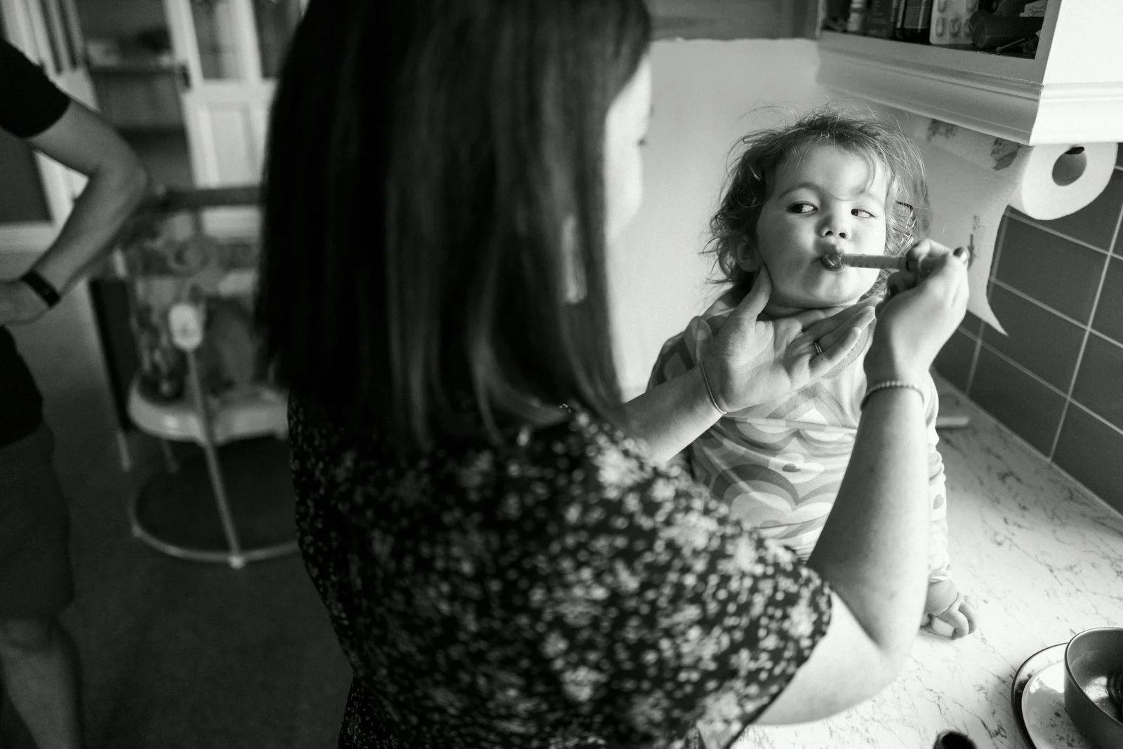 A woman helping a young girl eat a birthday cake with a candle, in a kitchen setting.