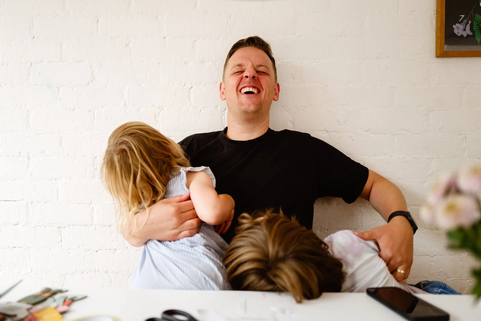A man laughing with two children, one with red hair and the other with brown hair, hugging him against a white brick wall.