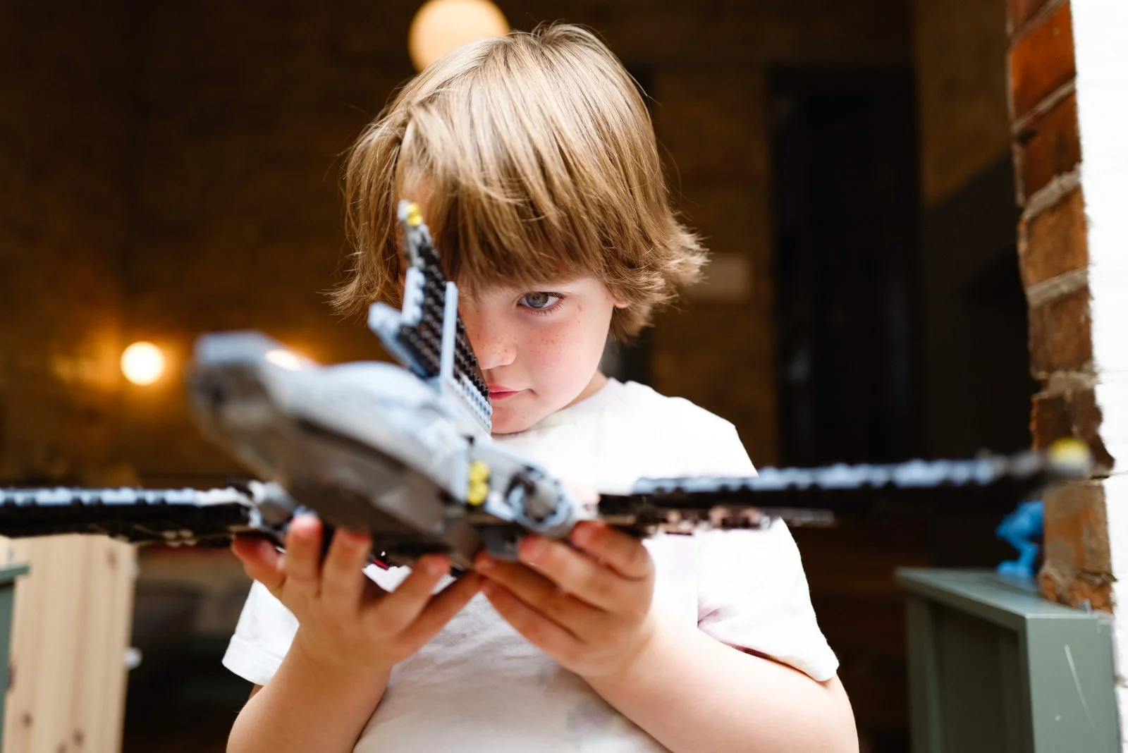 Young boy with light brown hair and freckles carefully examines a LEGO spaceship held upright, inside a cozy room with warm lighting and brick walls.