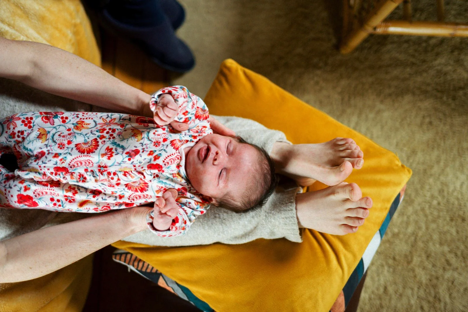 A newborn baby lying on a person's lap on an orange cushion, wearing a colorful floral outfit, with eyes closed and mouth open.