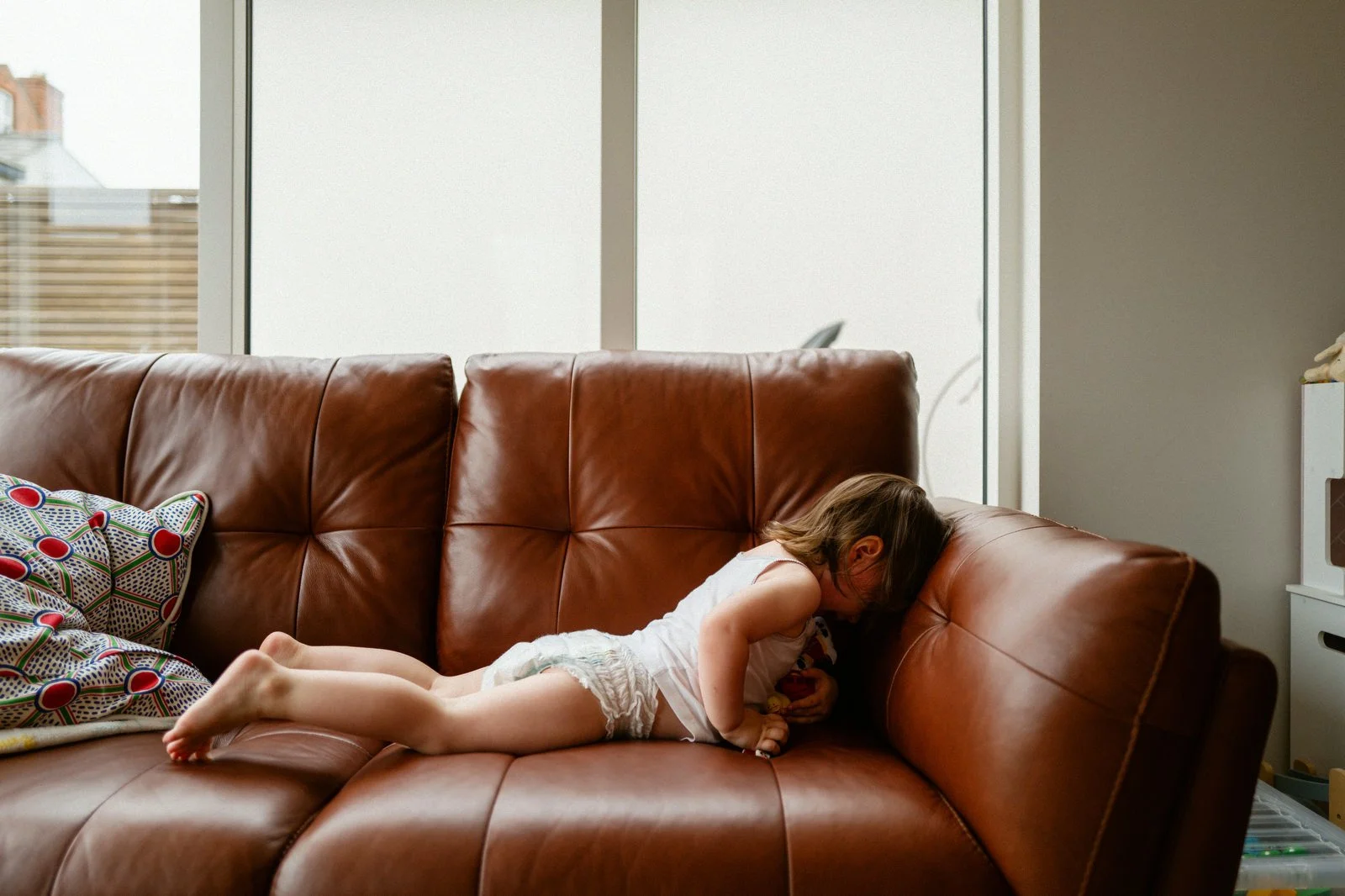 A young girl is lying on her stomach on a brown leather couch, with her head resting against the armrest and facing downward.