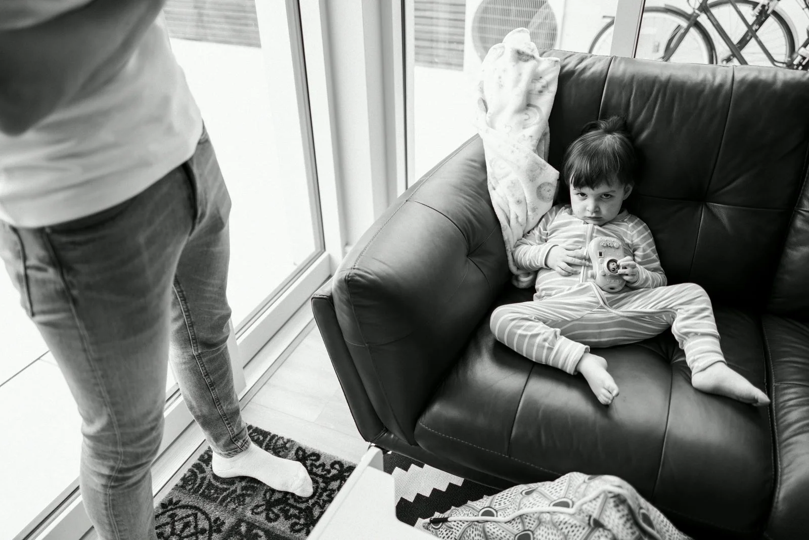 A young boy in striped pajamas lying on a black leather couch, holding a toy camera, with a blanket behind his head, while a person stands nearby near a glass door with an indoor bike visible outside.