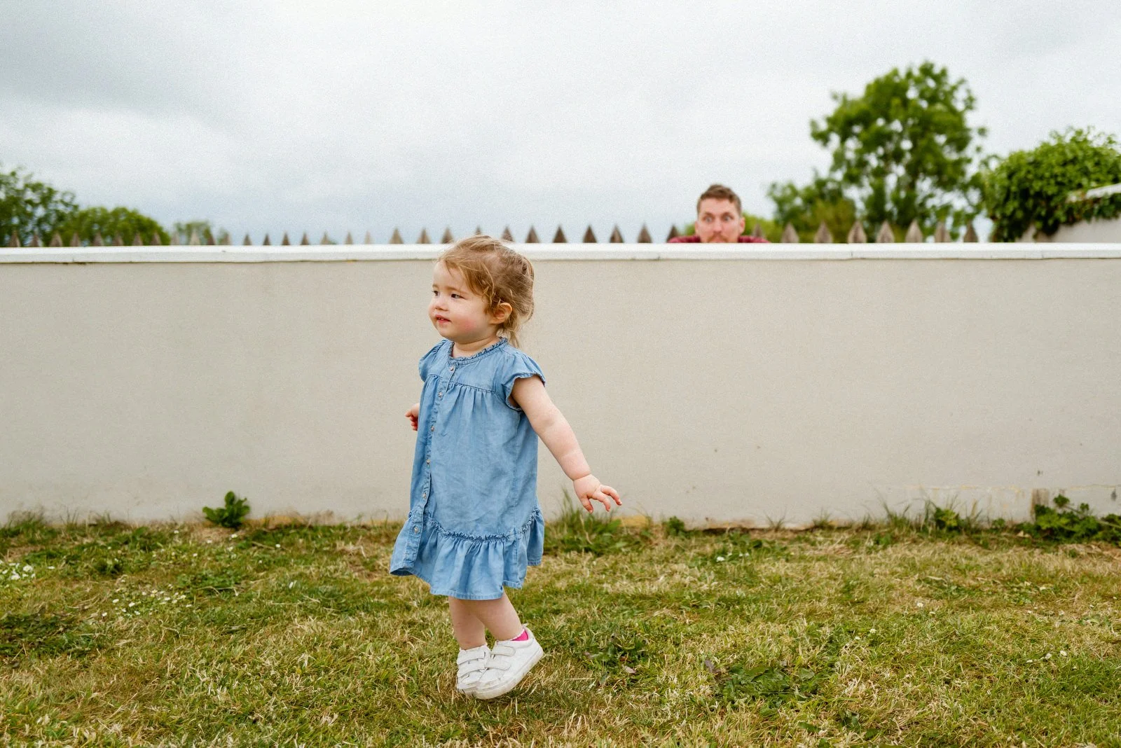 A young girl in a denim dress and white shoes walking on grass in a backyard, with a man peeking over a white wall behind her on a cloudy day.