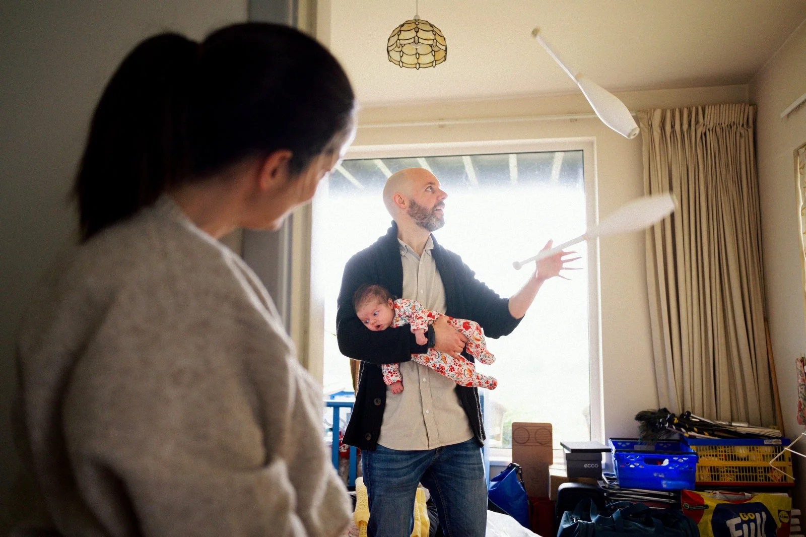 A man holding a baby inside a room, juggling with flying clubs, while a woman looks on. The room has a large window with curtains and various storage items.