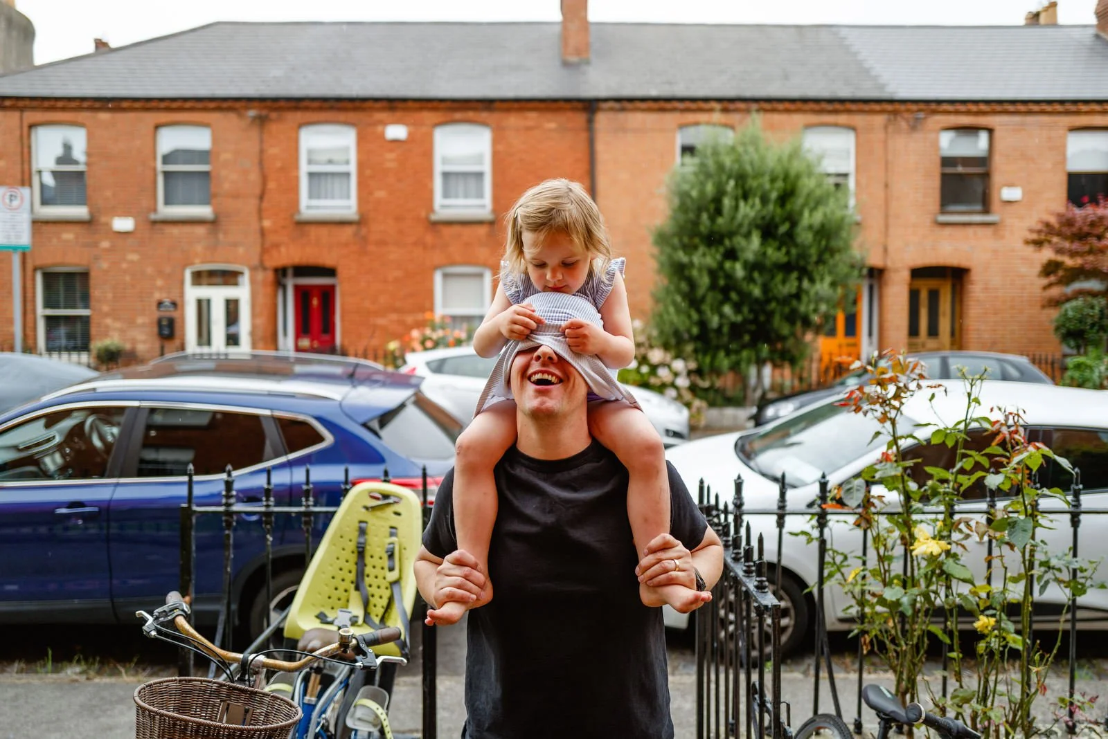 A man with a young girl sitting on his shoulders, covering his eyes with a blindfold, in a residential street with parked cars and brick houses.