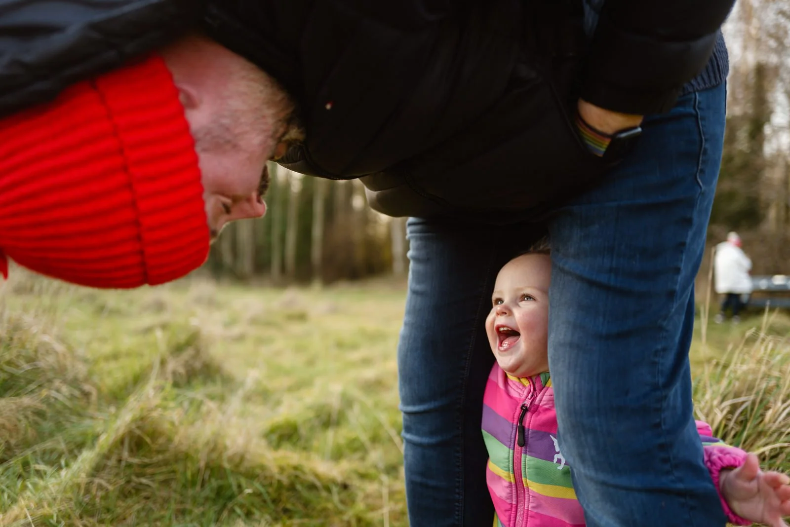 A young girl with a pink, purple, green, and yellow jacket laughing while standing underneath an adult, who is bent over at the waist and holding the girl. The adult's face is close to the girl's face, and the setting is an outdoor grassy area with t
