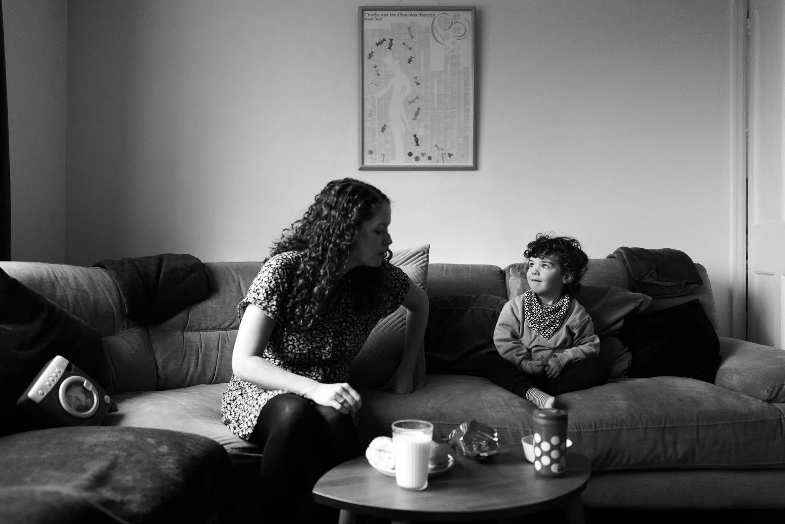 A woman and a young girl sitting on a living room sofa, engaged in a conversation. There is a coffee table with drinks and snacks in front of them, and a framed picture hanging on the wall behind them.