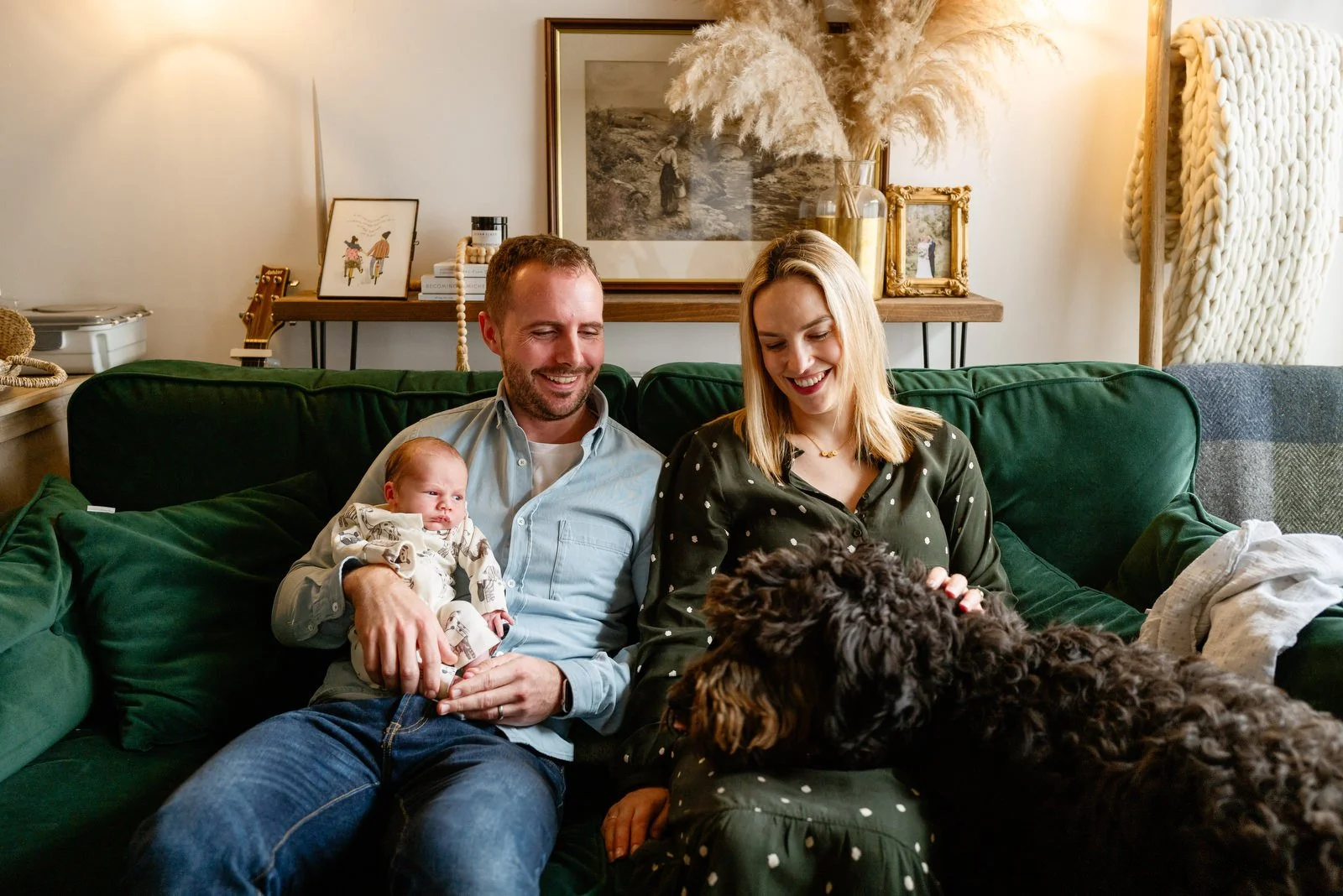 A happy family on a green couch with a baby and a black dog in a cozy, decorated living room.