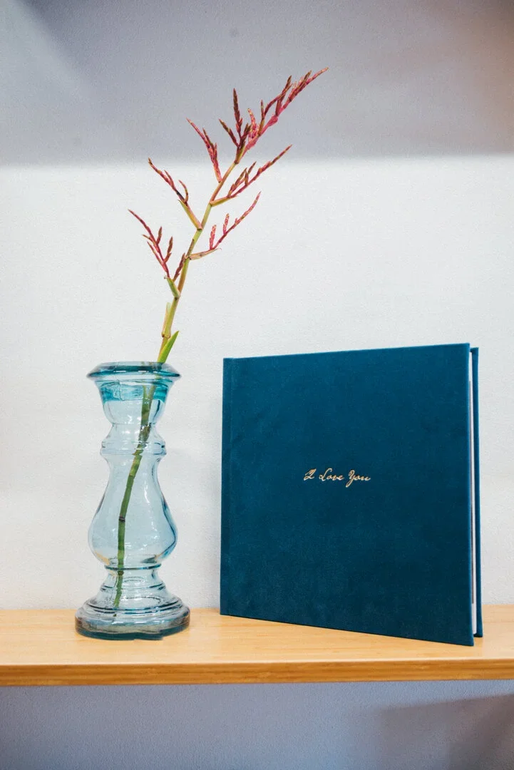 A clear glass vase with a single stem of red-tipped pinkish plant next to a blue booklet with gold lettering that says 'I love you' on a wooden shelf, against a plain wall background.