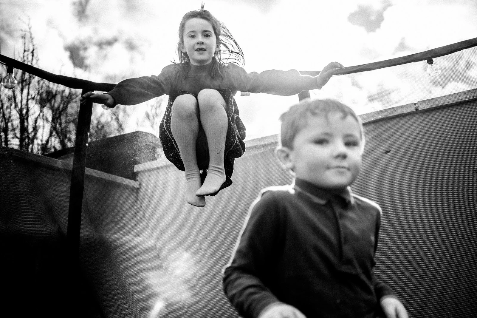 A girl sitting on a trampoline's edge, with her legs crossed and hands on the safety bar, mid-air, while a boy stands nearby on the ground, both outdoors with trees and a cloudy sky in the background.