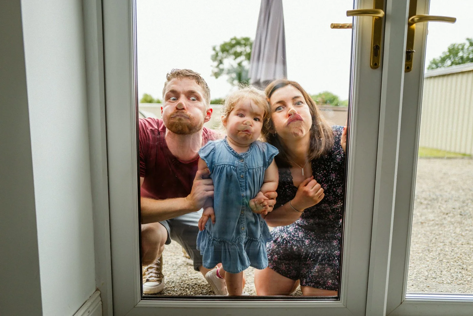 Three people, a man, a woman, and a young girl, making funny faces while looking through a glass door from outside.