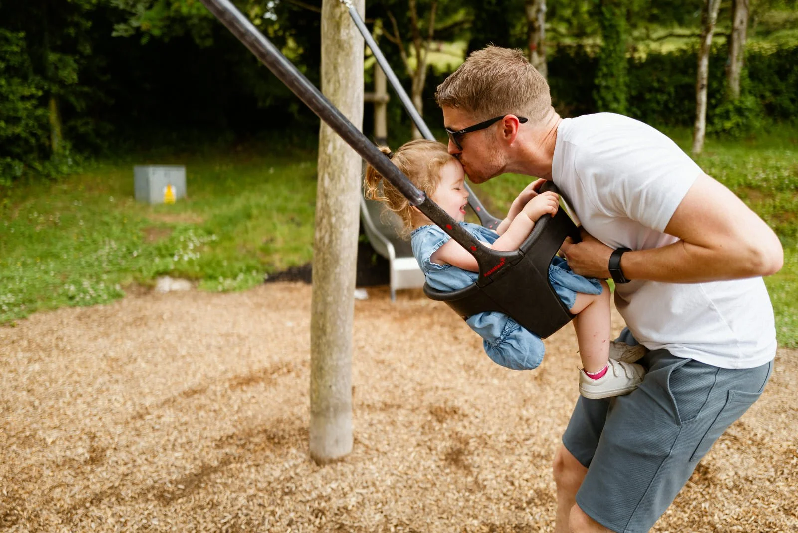 A man is bending down to kiss a young girl on the cheek while she is sitting in a swing at a playground. The girl is smiling and the man is wearing sunglasses and a white t-shirt. The background features trees and other playground equipment.