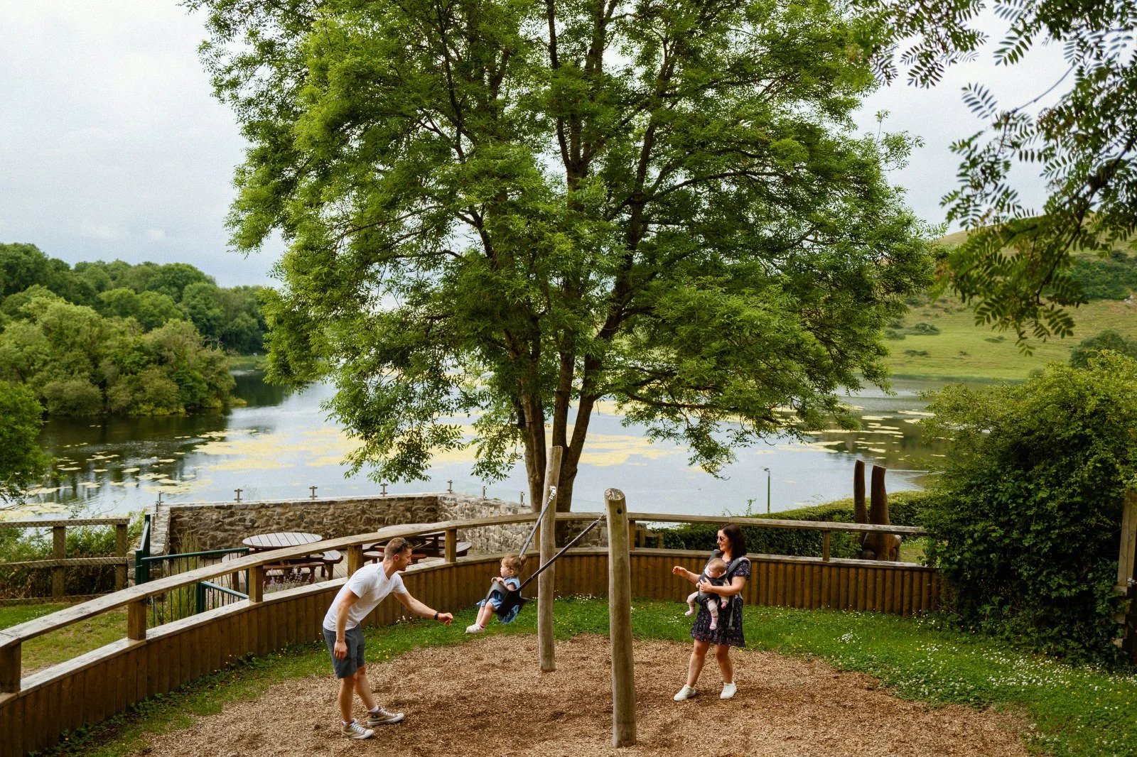 A family of four enjoying time together in a park with a pond and large tree in the background. The father is pushing a child on a swing, and the mother is holding another child, watching and smiling.