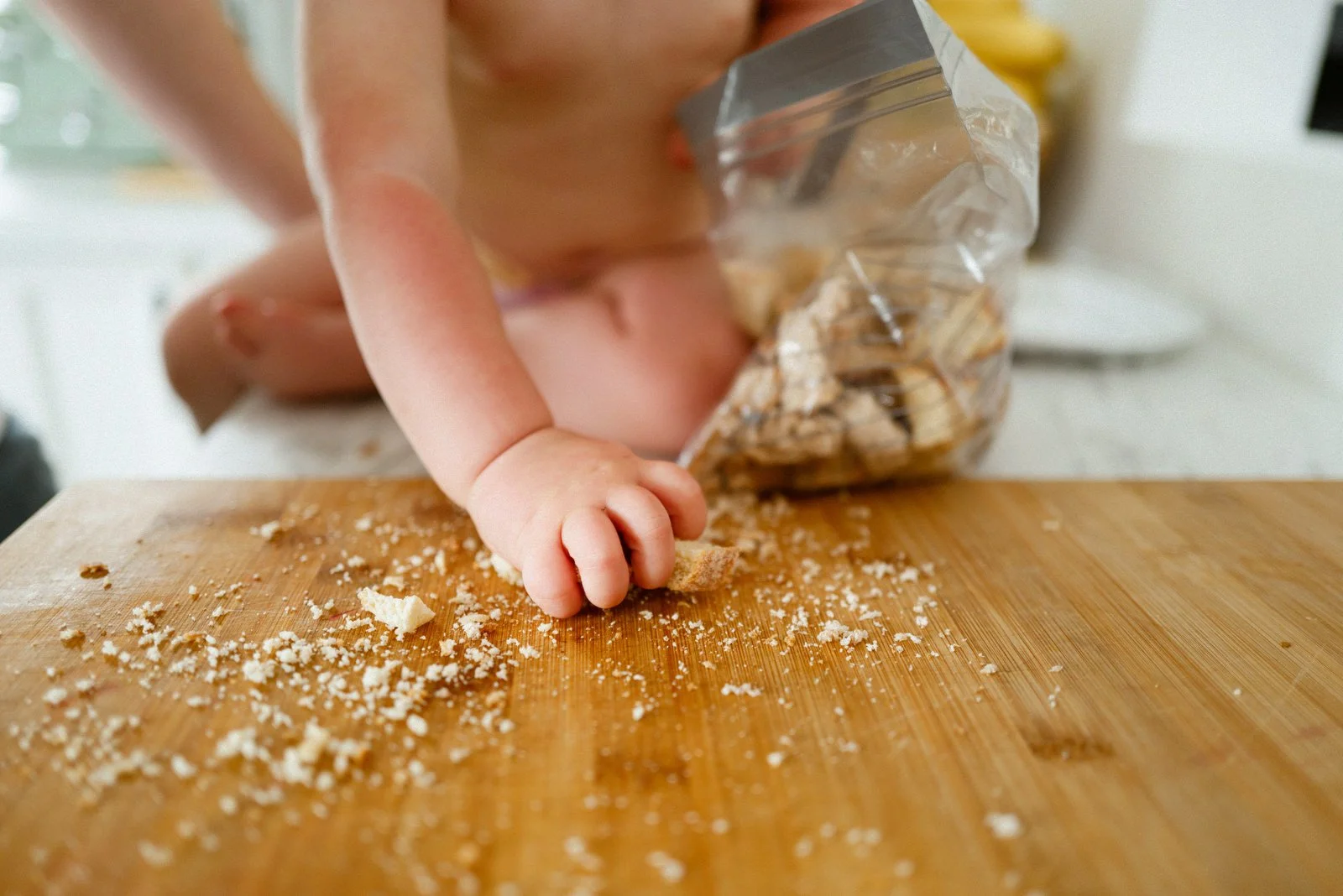 A young child's hand reaching for crumbs on a wooden cutting board, with a bag of snacks in the background.