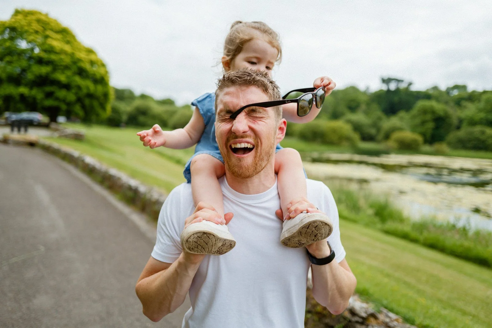 A man with a beard carrying a young girl on his shoulders outdoors near a pond with greenery. The girl is holding sunglasses and playing with the man's face, with a joyful expression. The man appears to be smiling and enjoying the moment.
