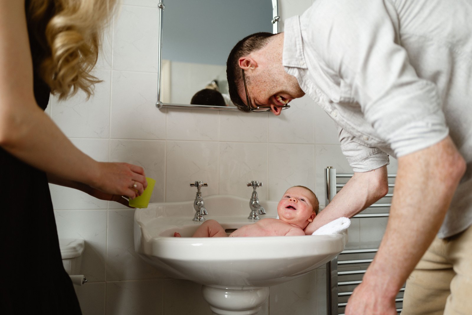 A baby being bathed in a white sink by two adults in a bathroom.
