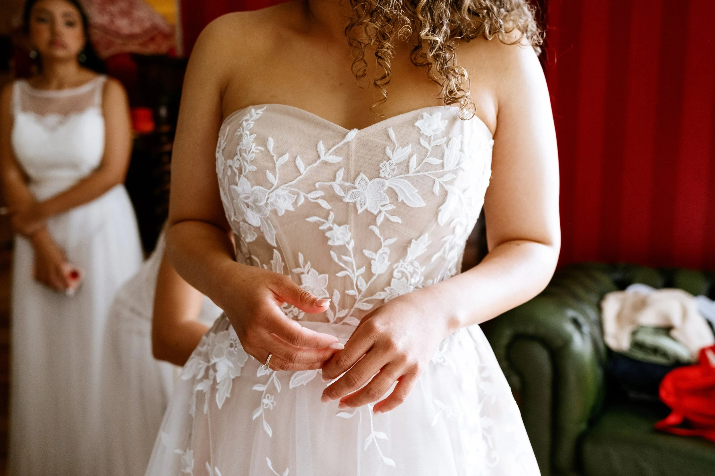 A bride in a strapless wedding dress with floral embroidery, standing with her hands clasped. In the background, another woman in a white dress is seen, with a green couch and red curtains behind them.