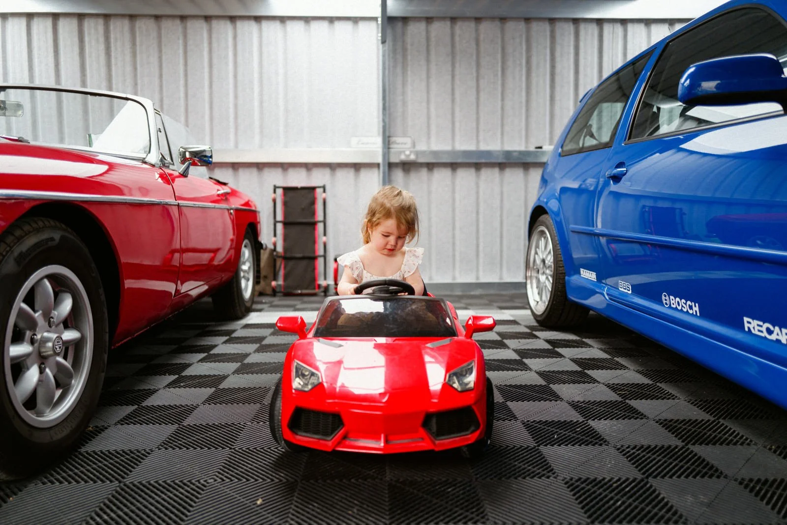 A young girl sitting in a red toy sports car parked between two real cars, one red and one blue, in a garage with corrugated metal walls.