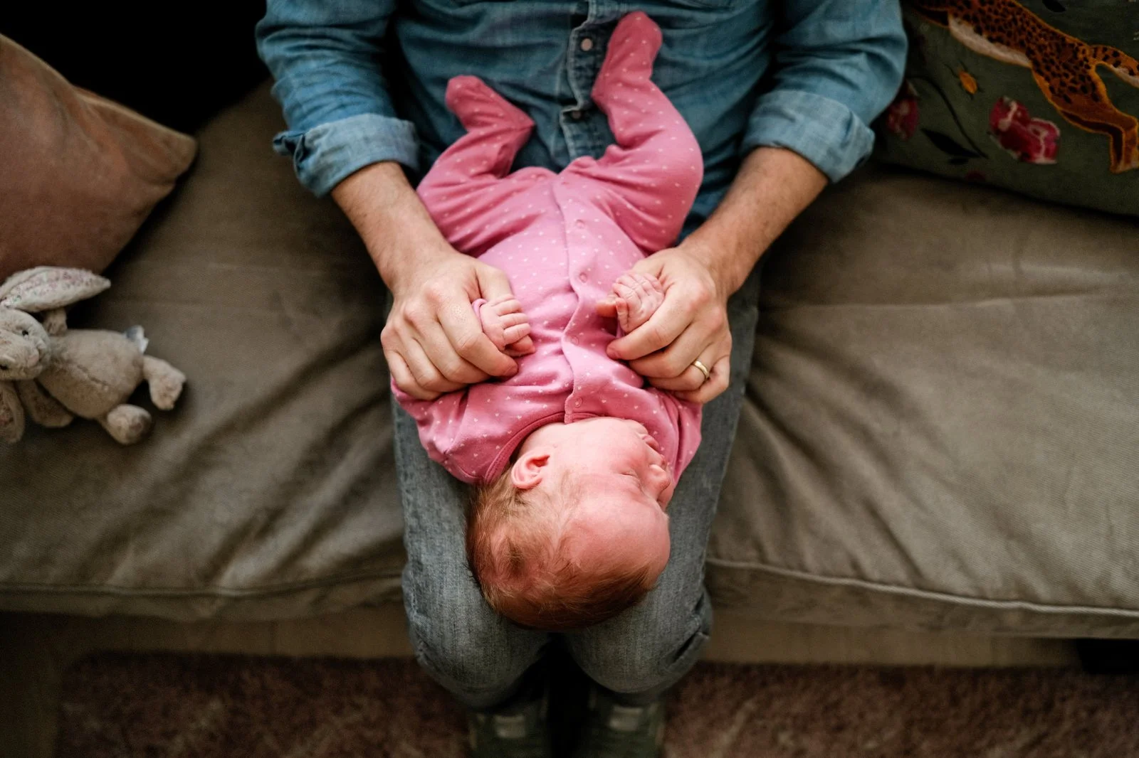 A person in a denim shirt holding a newborn baby dressed in pink on their lap, sitting on a beige sofa.