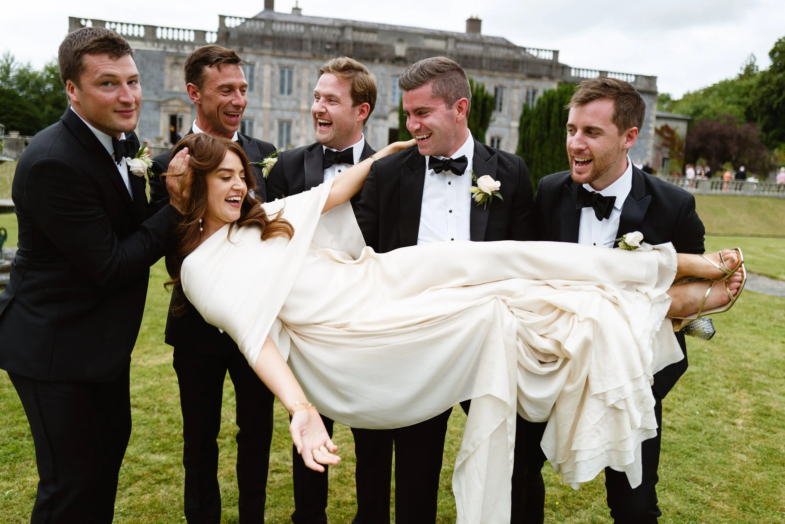 Groom and bridesmaids holding a bride in a white dress, smiling and laughing outdoors with a large stone building in the background.