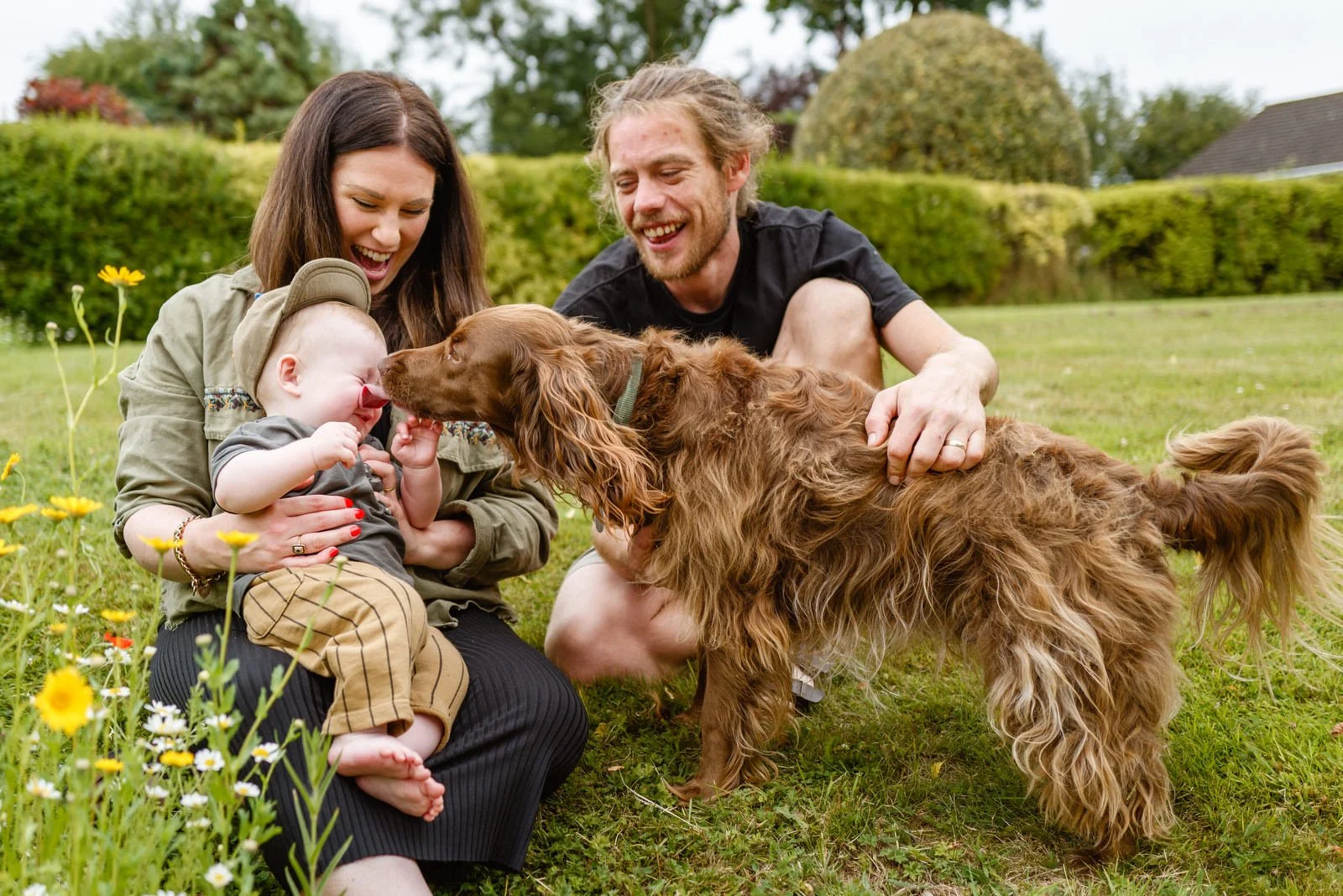 A family of three, including a woman, a man, a young child, and a dog, sitting on a grassy lawn with flowers and a hedge in the background. The woman and the dog are touching noses with the child smiling.