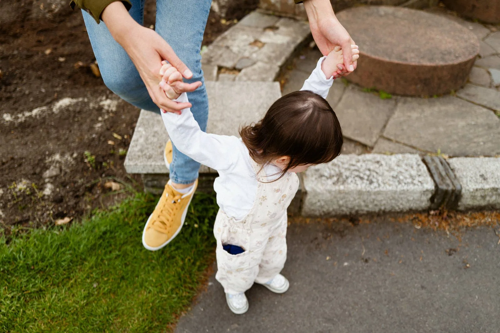 A young child with brown hair, wearing white overalls and white shoes, holding hands with an adult while standing on a sidewalk near a flower bed and grass.