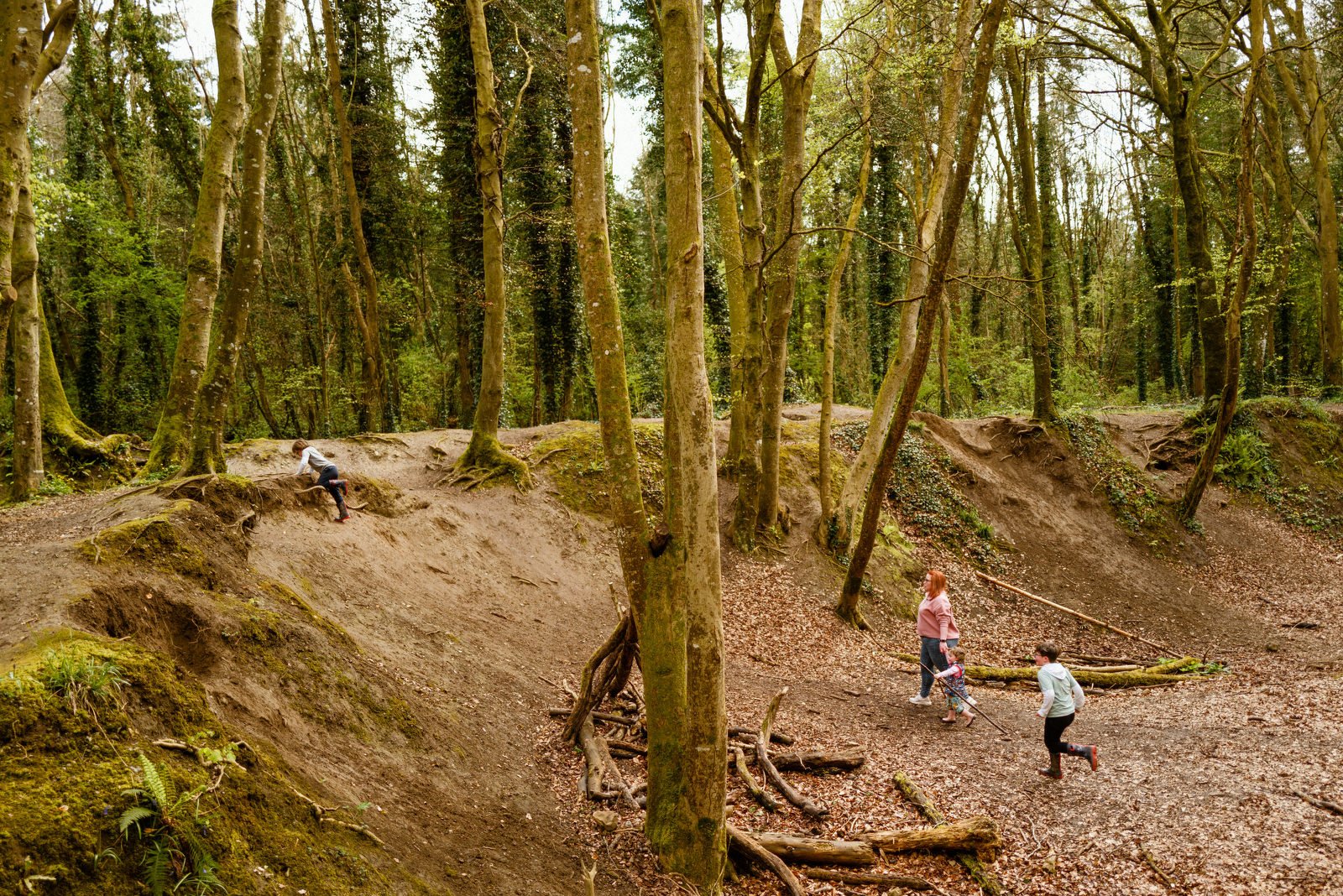 A woman and two children walking through a forest, one child climbing a dirt hill and another running with a stick.