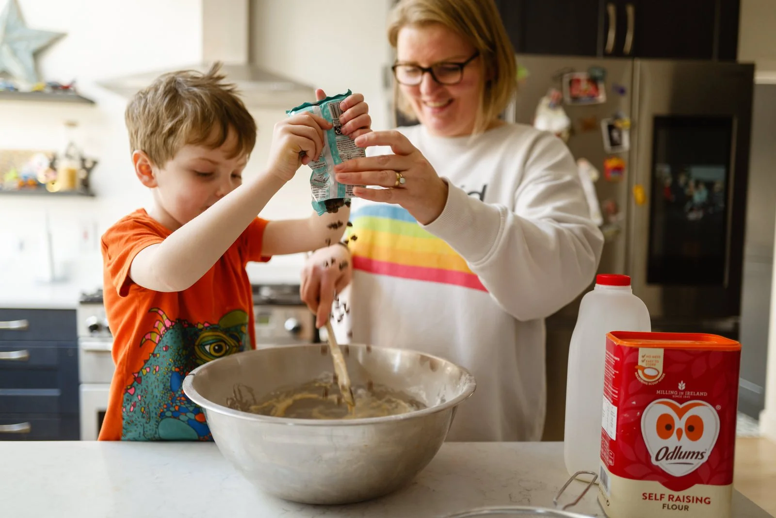 A woman and a young boy baking together in a kitchen. The boy is pouring ingredients from a bag into a large mixing bowl while the woman helps and smiles. Kitchen items and a refrigerator are visible in the background.